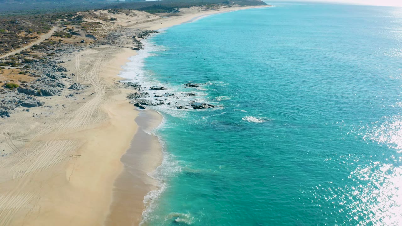 Aerial view over La Fortuna, Baja California Sur, with turquoise ocean beside pale sand, scattered dark rocks, inland dunes, tire tracks, and no visible people.