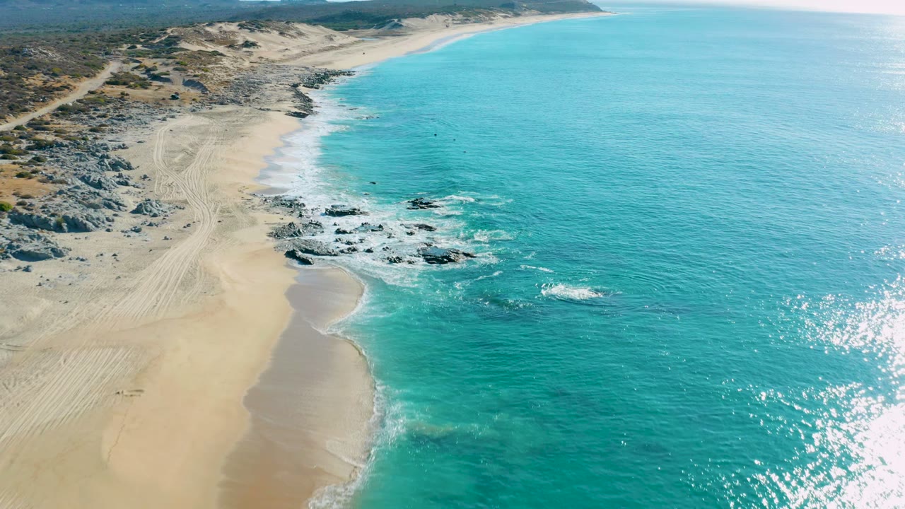 Turquoise surf breaks near dark rocks along a wide sandy shoreline.