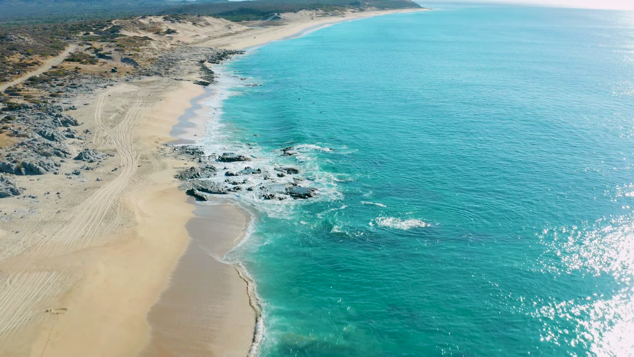 Aerial view at La Fortuna with the shoreline curving past pale beach sand, dark rocks in the surf, bright turquoise water, dune terrain, and no visible people.
