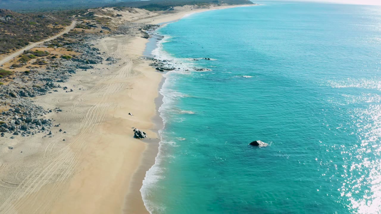 Aerial frame over La Fortuna beach in Baja California Sur showing pale sand, turquoise water, rocky shallows, tire tracks, and no visible people.