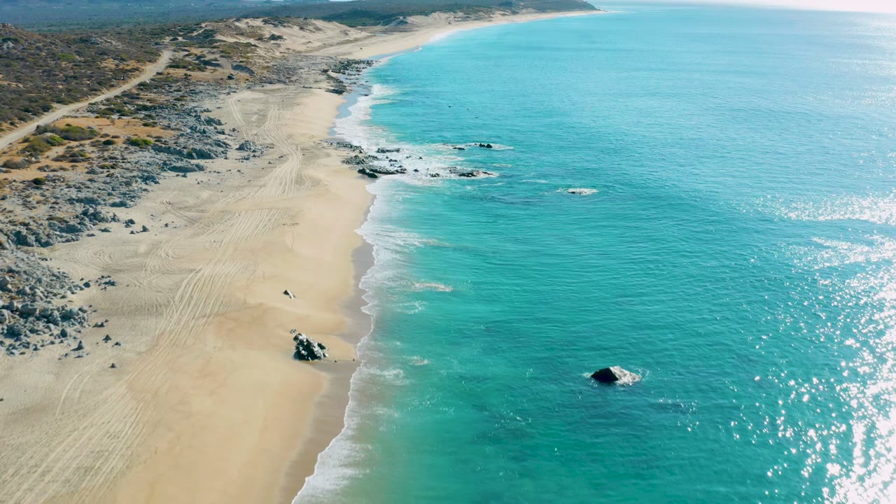 Drone view of turquoise shoreline with beach tracks and a small dark vehicle on sand.