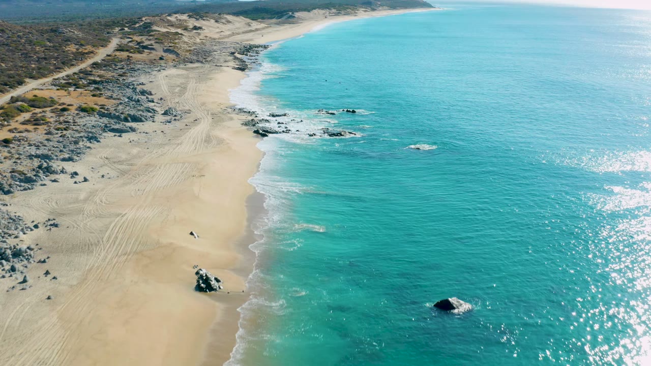 Aerial frame along La Fortuna's shoreline showing the beach curving past rock clusters into clear turquoise water, with no visible people.