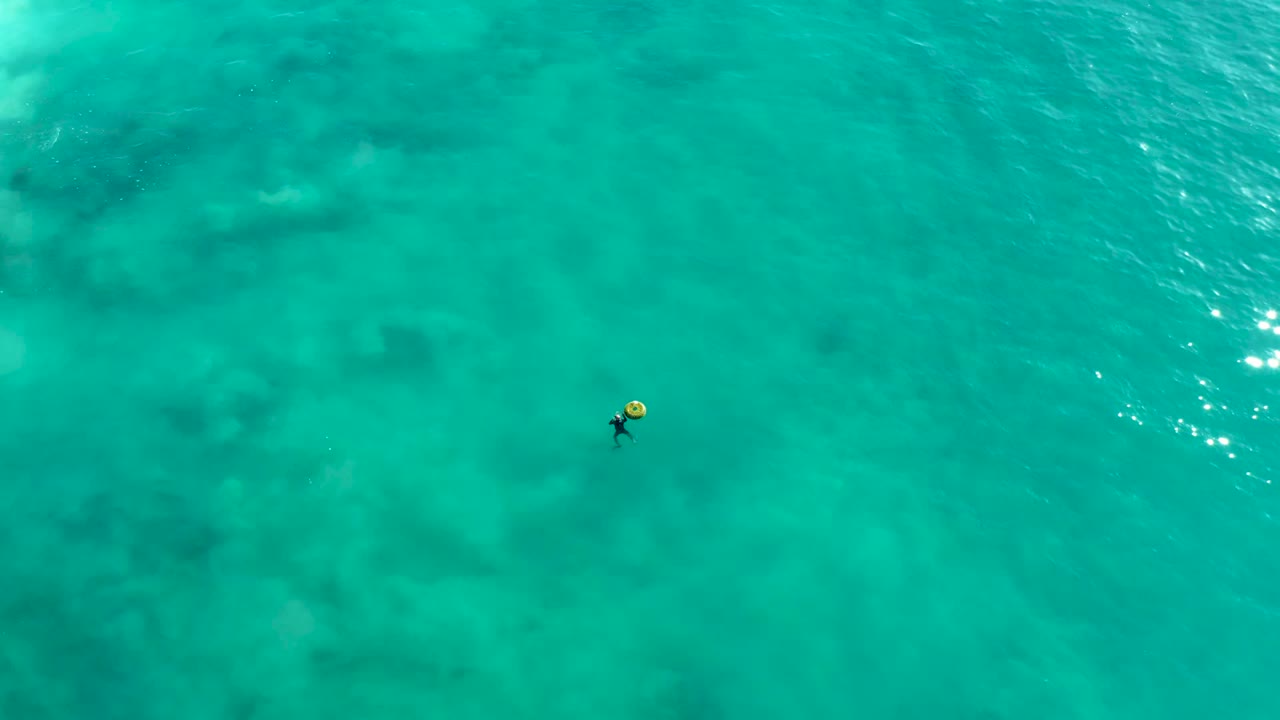 At La Fortuna, Baja California Sur, an overhead view shows one person with a yellow float near the left side of clear turquoise ocean water with darker submerged patches.