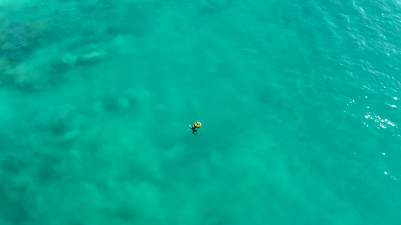 At La Fortuna, Baja California Sur, the swimmer remains small in the overhead frame while darker underwater rock or reef shapes fill the upper part of the turquoise water.