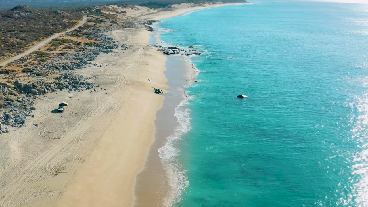 Aerial frame over La Fortuna, Baja California Sur, showing pale sand, turquoise water, rocky shoreline, vehicle tracks, a small tent-like object on the beach, and no visible people.