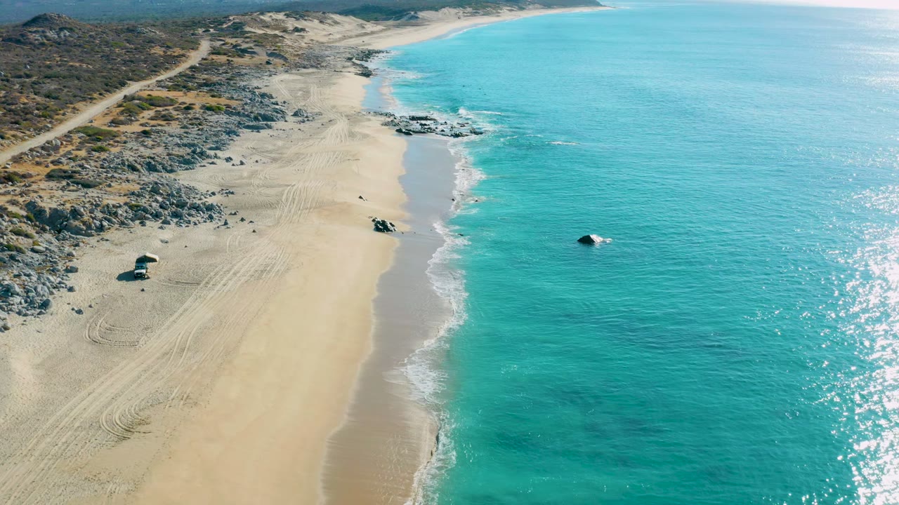 Drone view of turquoise water beside a sandy shore with rocks, tracks, and a small beach object.