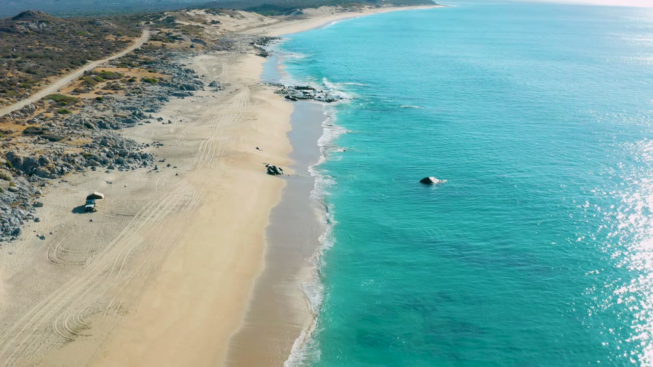 High drone frame over La Fortuna, Baja California Sur, with rocky outcrops filling the upper beach, open turquoise water on the right, sand tracks below, and no visible people.