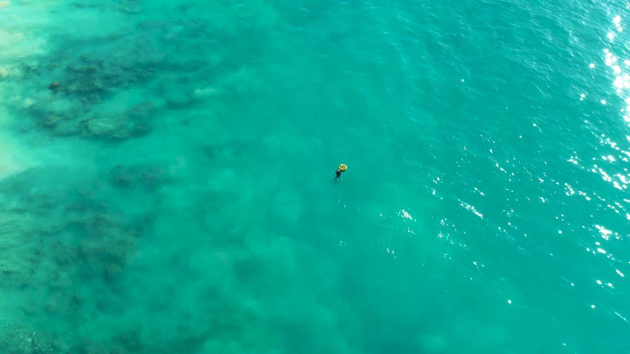 Aerial view over La Fortuna turquoise ocean water with a small snorkeler near the left center, dark seabed patches below, and bright surface reflections.