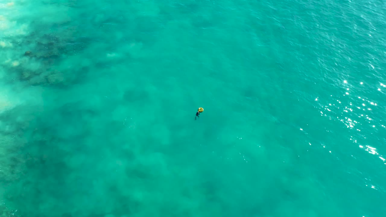 Aerial view at La Fortuna with a small snorkeler toward the upper right, bright sun glints on the water, and darker seabed forms visible beneath the turquoise surface.