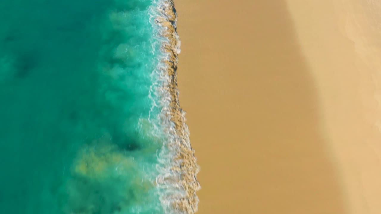 Overhead view at La Fortuna, Baja California Sur, Mexico, showing turquoise ocean on the left, a narrow white surf edge, pale sand on the right, and no visible people.
