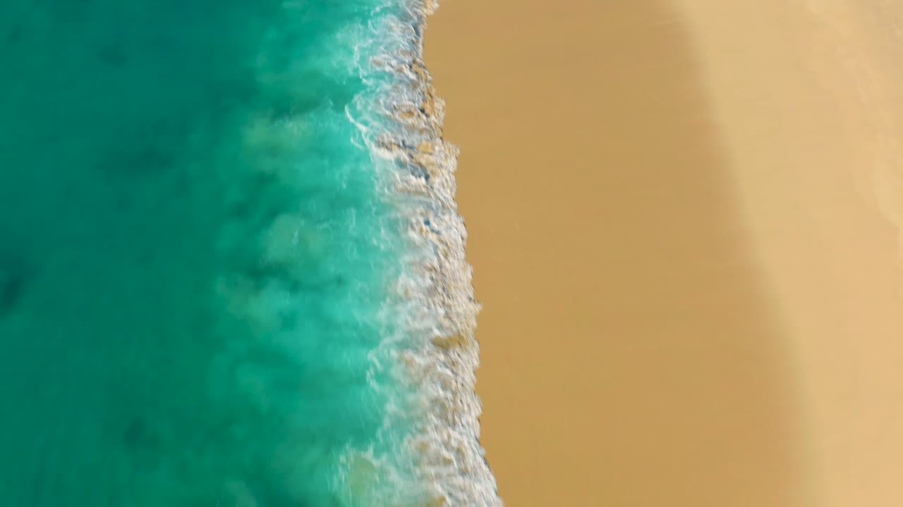 Aerial shoreline with turquoise water, breaking foam, and smooth sand.