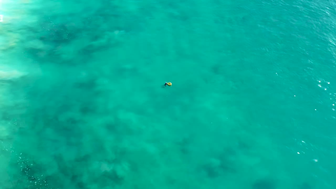Overhead view in La Fortuna, Baja California Sur, showing a tiny snorkeler with a yellow float near the center of clear turquoise water with darker underwater patches.