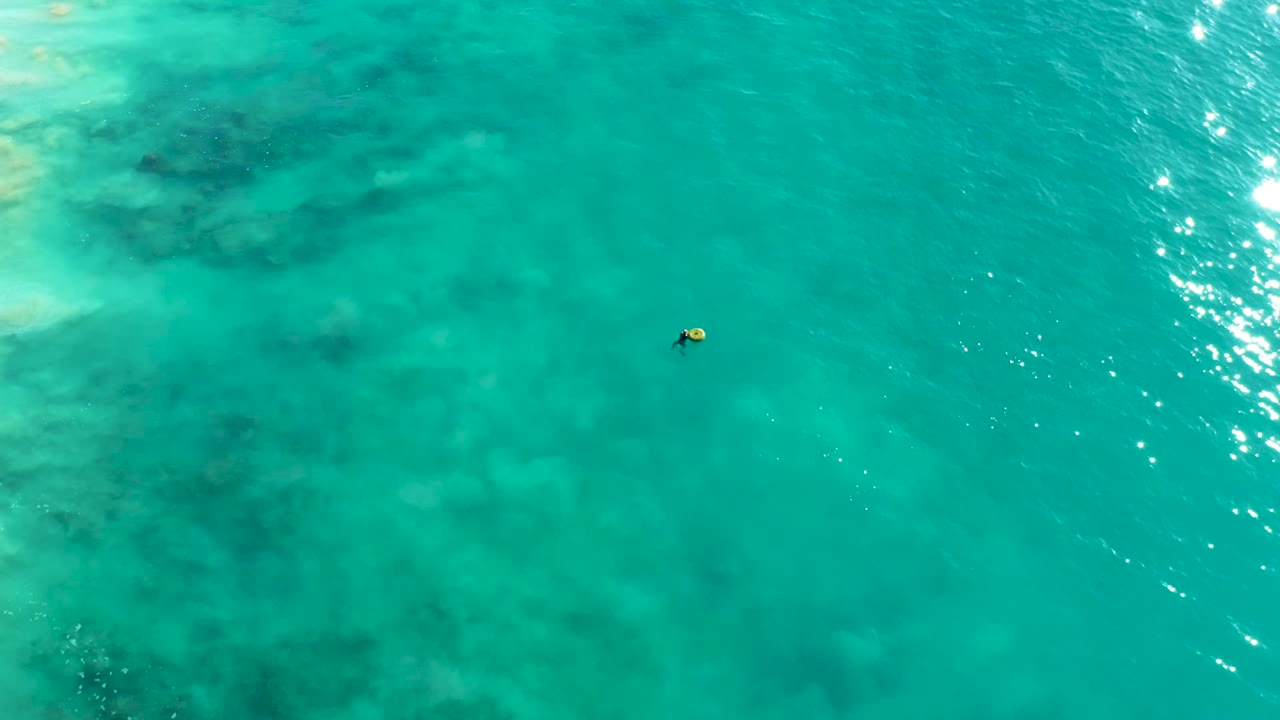 Overhead La Fortuna water view with the distant snorkeler and yellow float left of the bright sun reflections along the right side of the frame.