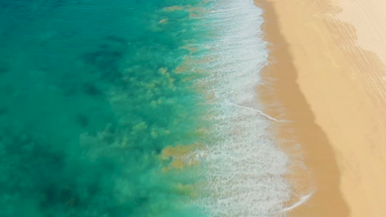 Overhead view of turquoise water washing onto pale sand in La Fortuna, Baja California Sur, with darker underwater patches and no visible people.