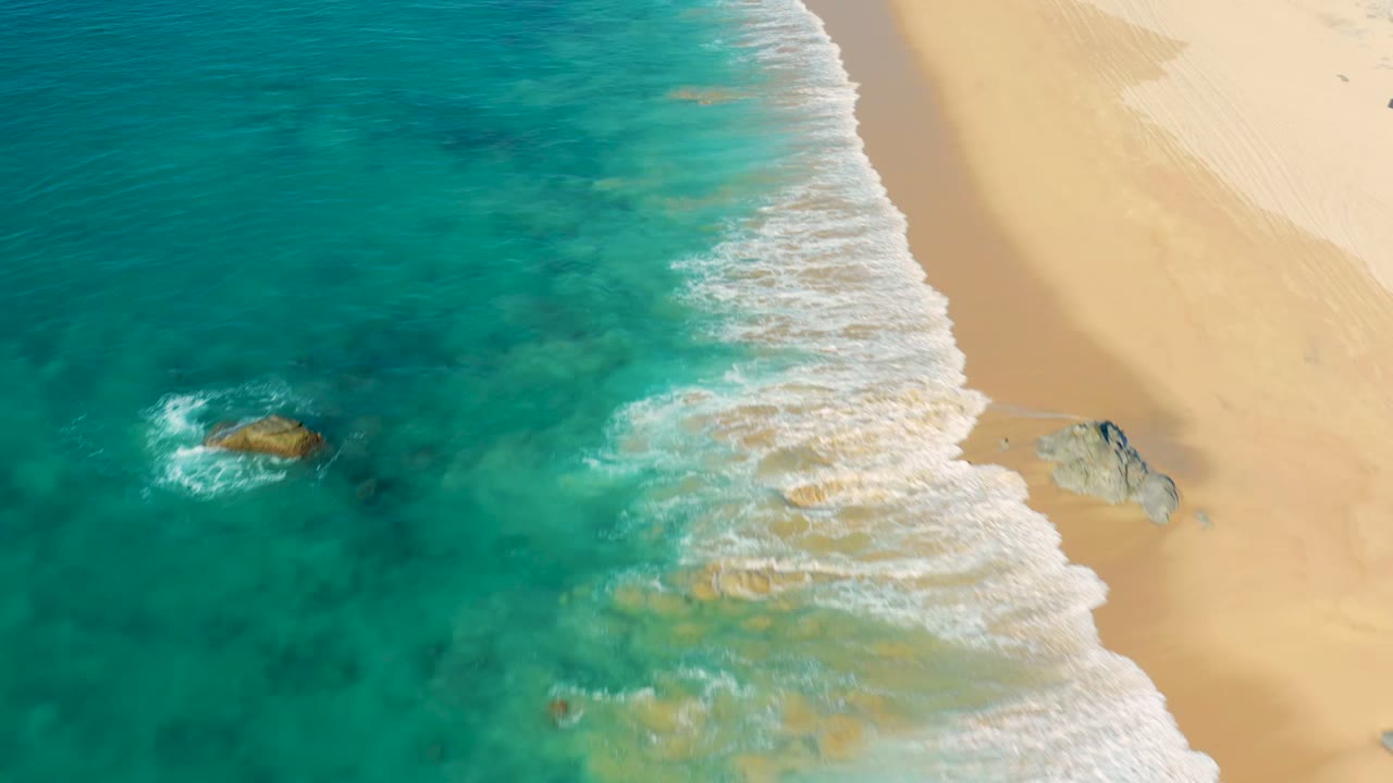 Aerial frame from La Fortuna, Baja California Sur, Mexico showing turquoise water on the left, white surf crossing the beach diagonally, pale sand on the right, scattered rocks, and no visible people.