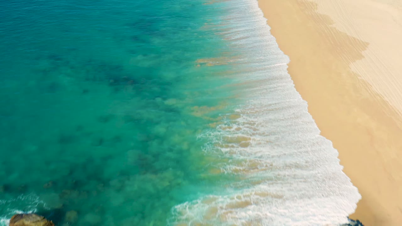Aerial frame from La Fortuna, Baja California Sur, Mexico showing the surf line shifted along the shore, with turquoise water, white foam, pale sand, partially cropped rocks, and no visible people.