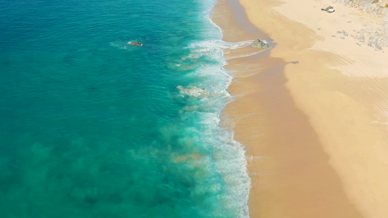 Aerial frame from La Fortuna, Baja California Sur showing turquoise ocean on the left, pale sand on the right, a small offshore rock, and no visible people.