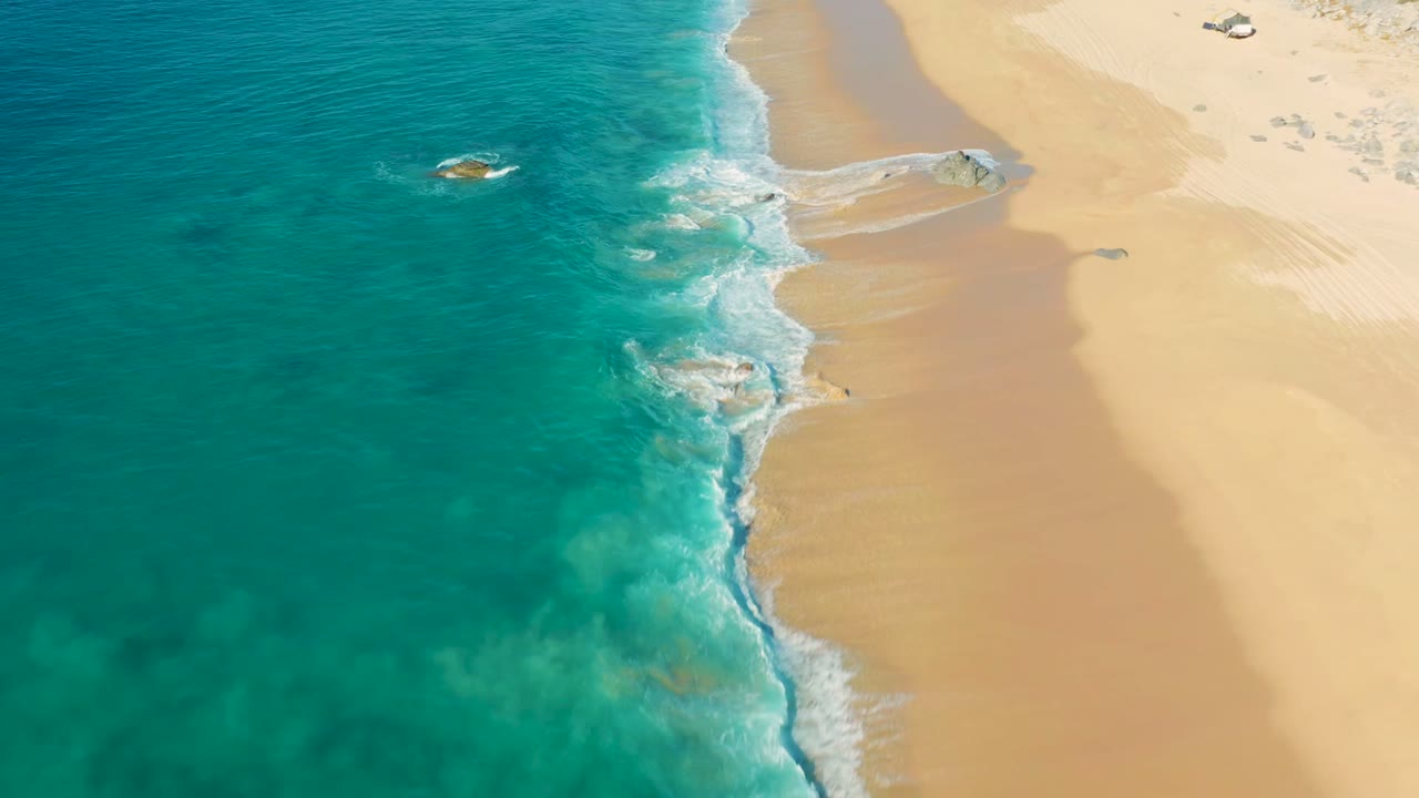 Turquoise surf reaches a pale sandy beach from an overhead angle.