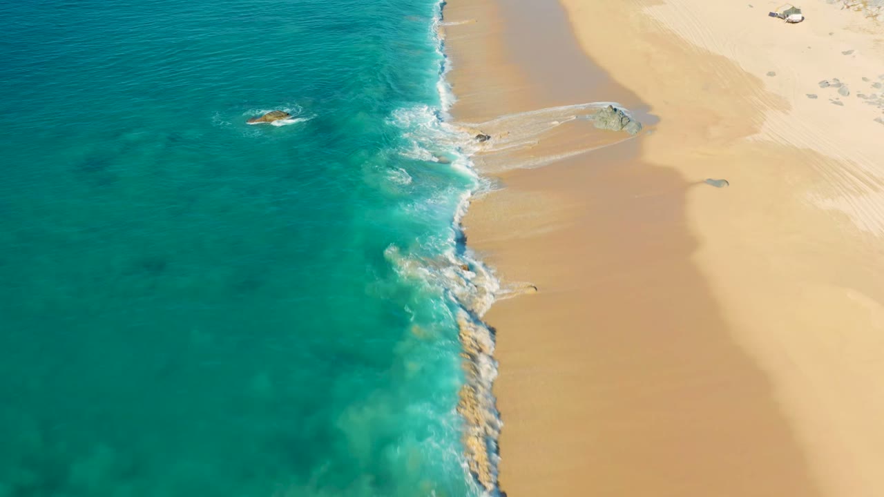 Aerial frame from La Fortuna, Baja California Sur where the surf line thickens around exposed rocks, with open sand to the right and no visible people.