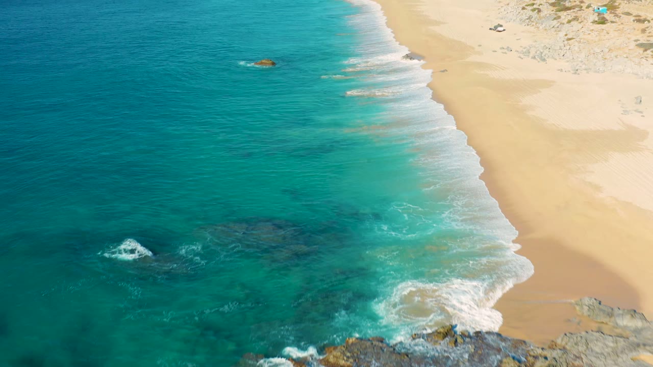 Aerial view at La Fortuna, Baja California Sur, with turquoise ocean on the left, pale sand on the right, white surf along the diagonal shoreline, rocks near the bottom edge, and no visible people.
