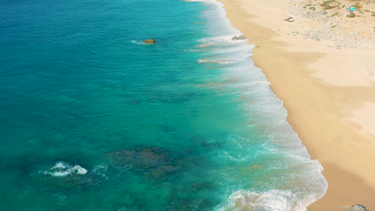 Turquoise sea and white waves beside a mostly empty sandy beach.