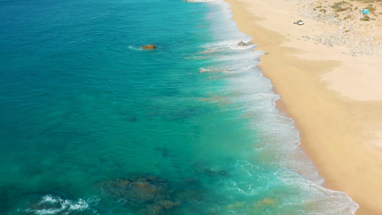 Aerial view over La Fortuna shoreline with broad turquoise water, darker reef-like rock patches under the surface, a narrow white surf edge, pale sand to the right, and no visible people.