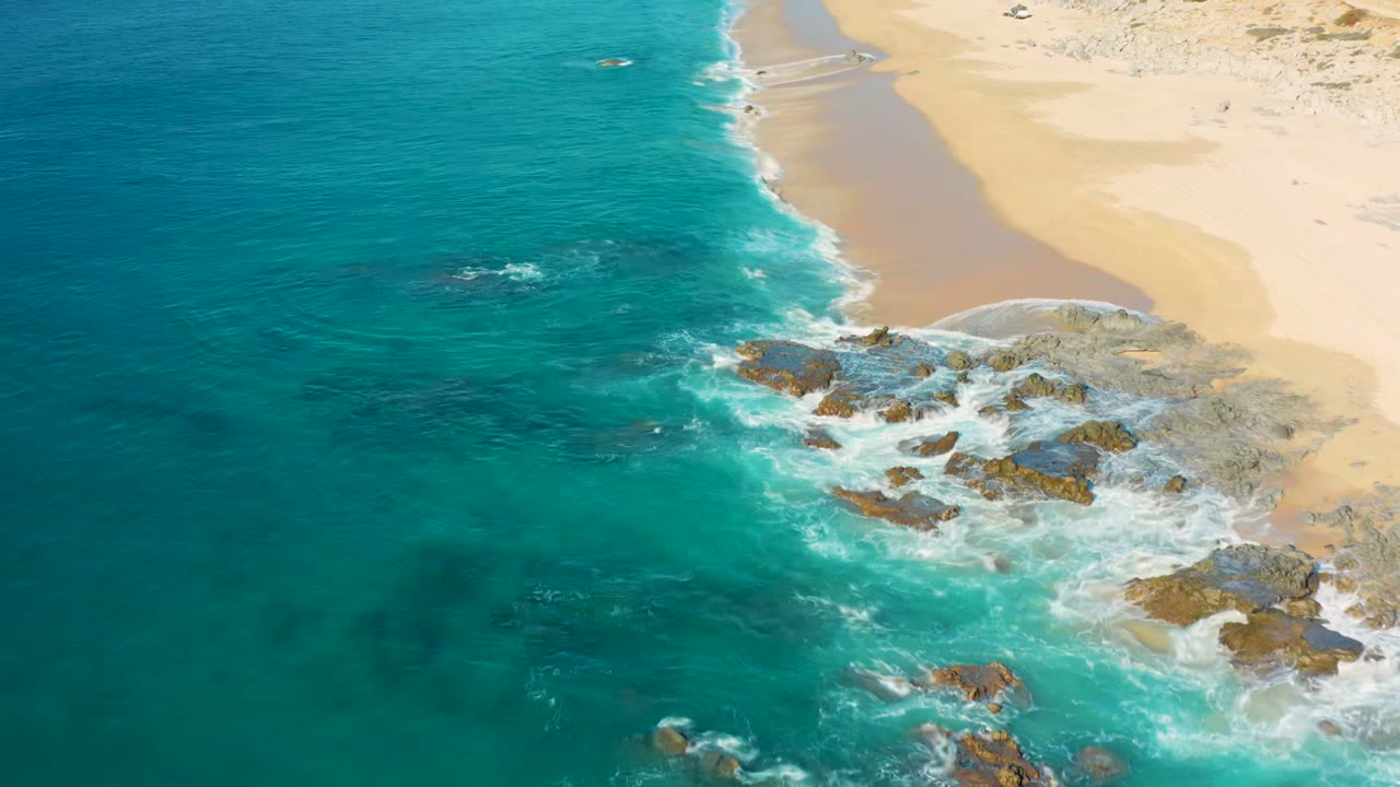Aerial view at La Fortuna, Baja California Sur, with turquoise ocean on the left, pale sand on the right, surf foaming around rocky outcrops, and no visible people.