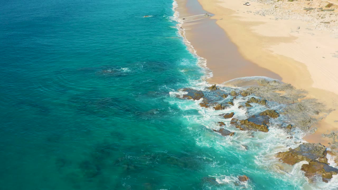 Turquoise surf curls around dark rocks along a pale sand beach.