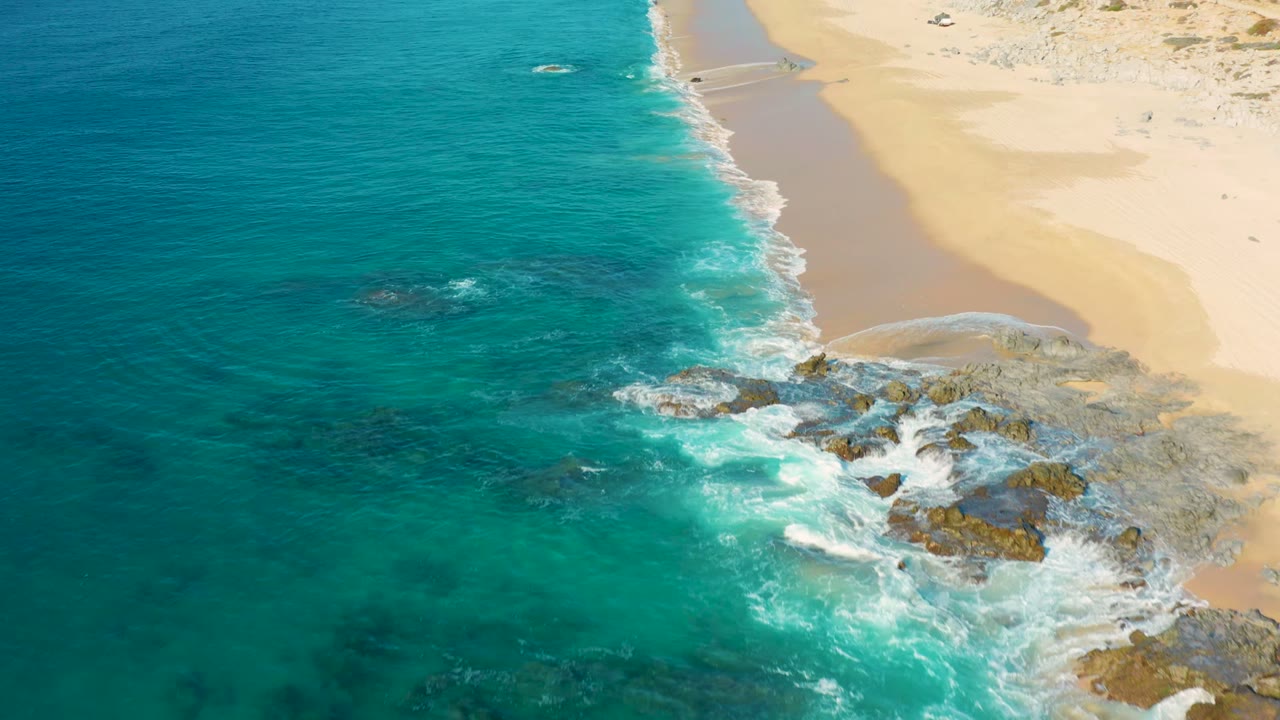 Aerial view of La Fortuna beach with the rocky surf line shifted toward the lower right, blue-green water at left, pale sand at upper right, and no visible people.