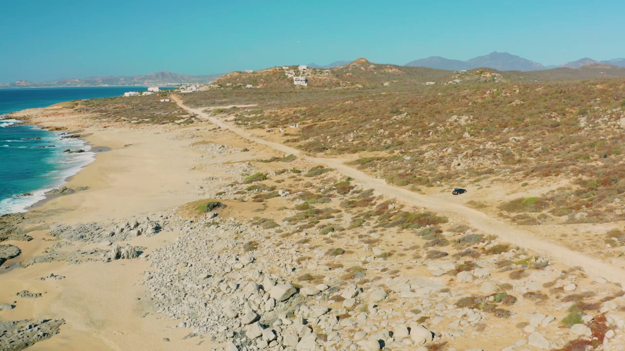 At 0:00, an aerial view in La Fortuna, Baja California Sur shows a dirt road crossing dry coastal scrub beside a sandy beach, turquoise ocean, rocks, distant hills, and no visible people.