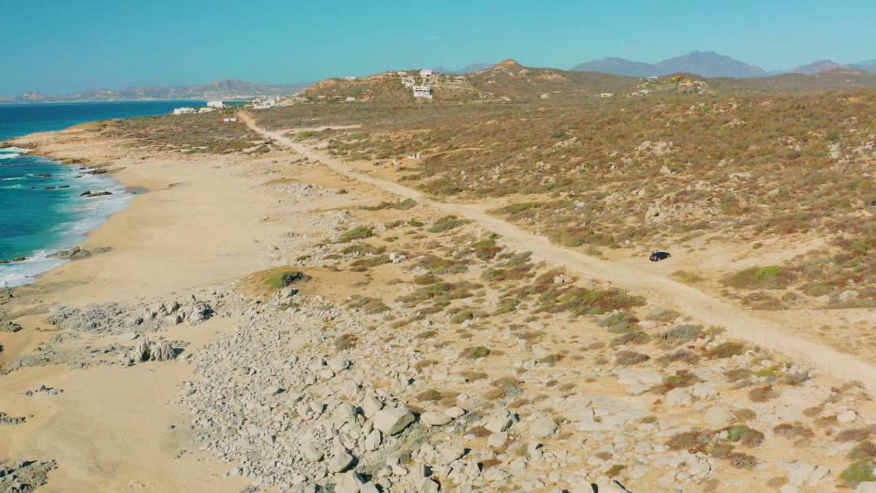At 0:02, the aerial composition in La Fortuna shows the dirt road cutting across arid scrubland above the beach, with the vehicle near the right side, waves on the left, distant hills, and no visible people.