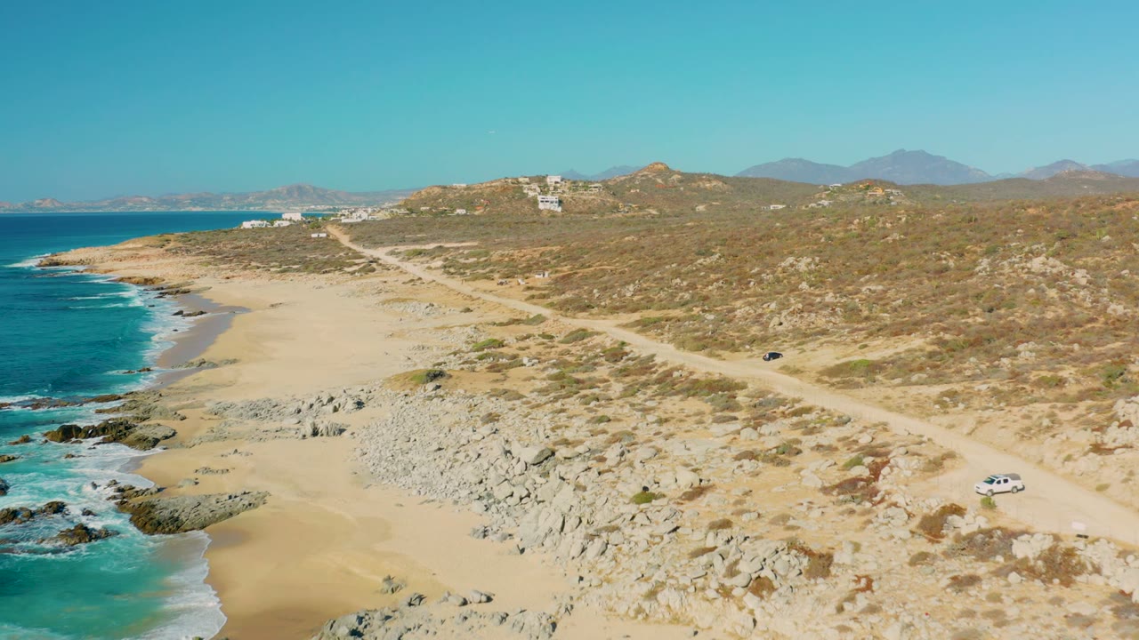 At 0:00, an aerial view in La Fortuna, Baja California Sur shows a white car on a dirt road above a sandy beach, turquoise surf, rocky foreground, dry hills, and no visible people.