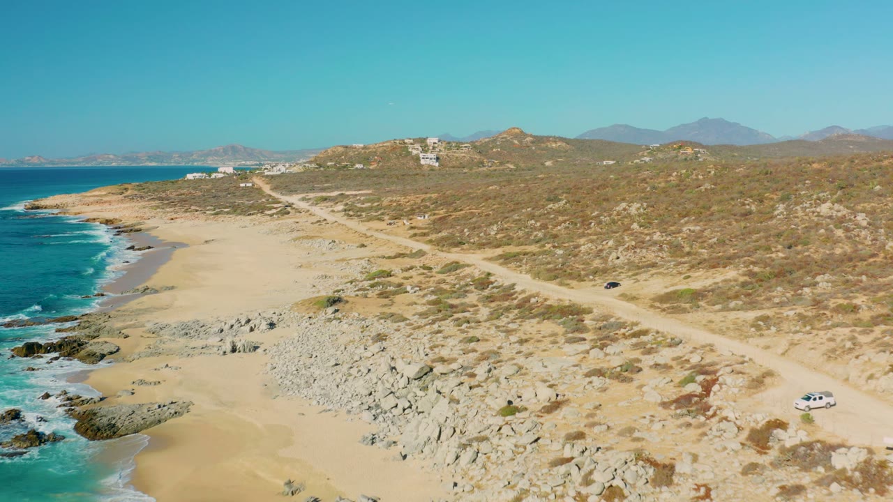 White car continues along a dirt road beside a sandy coast and rocky beach.