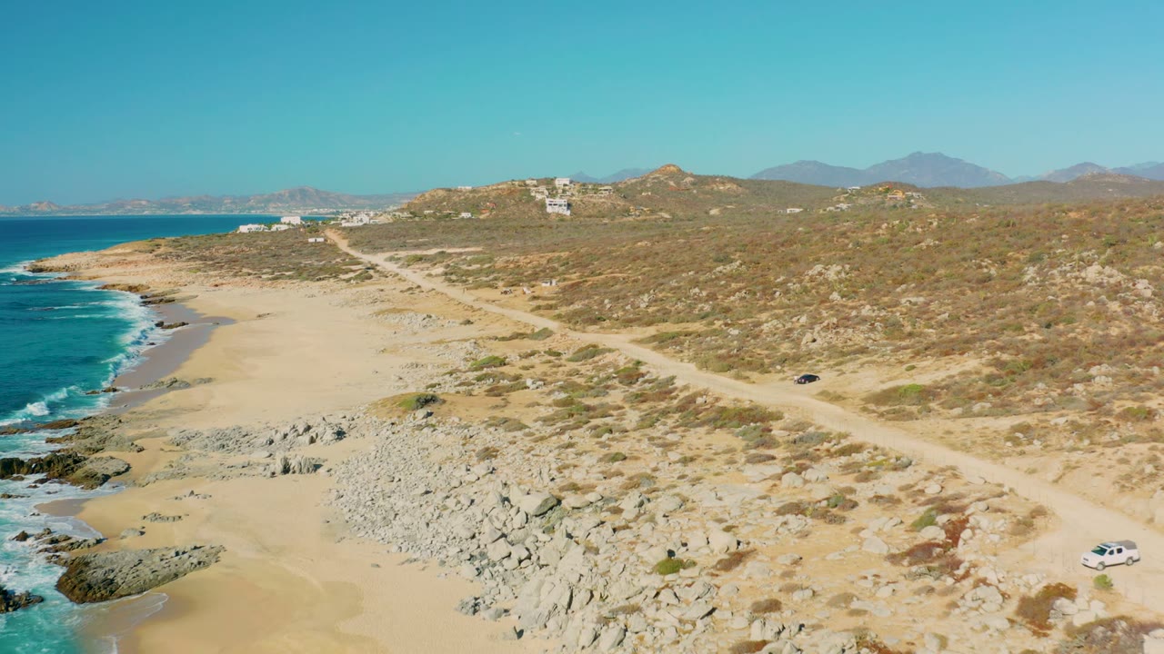 At 0:02, the car sits farther along the pale dirt road in La Fortuna, with the sandy beach curving below, rocky outcrops near the water, dry hillside, and no visible people.