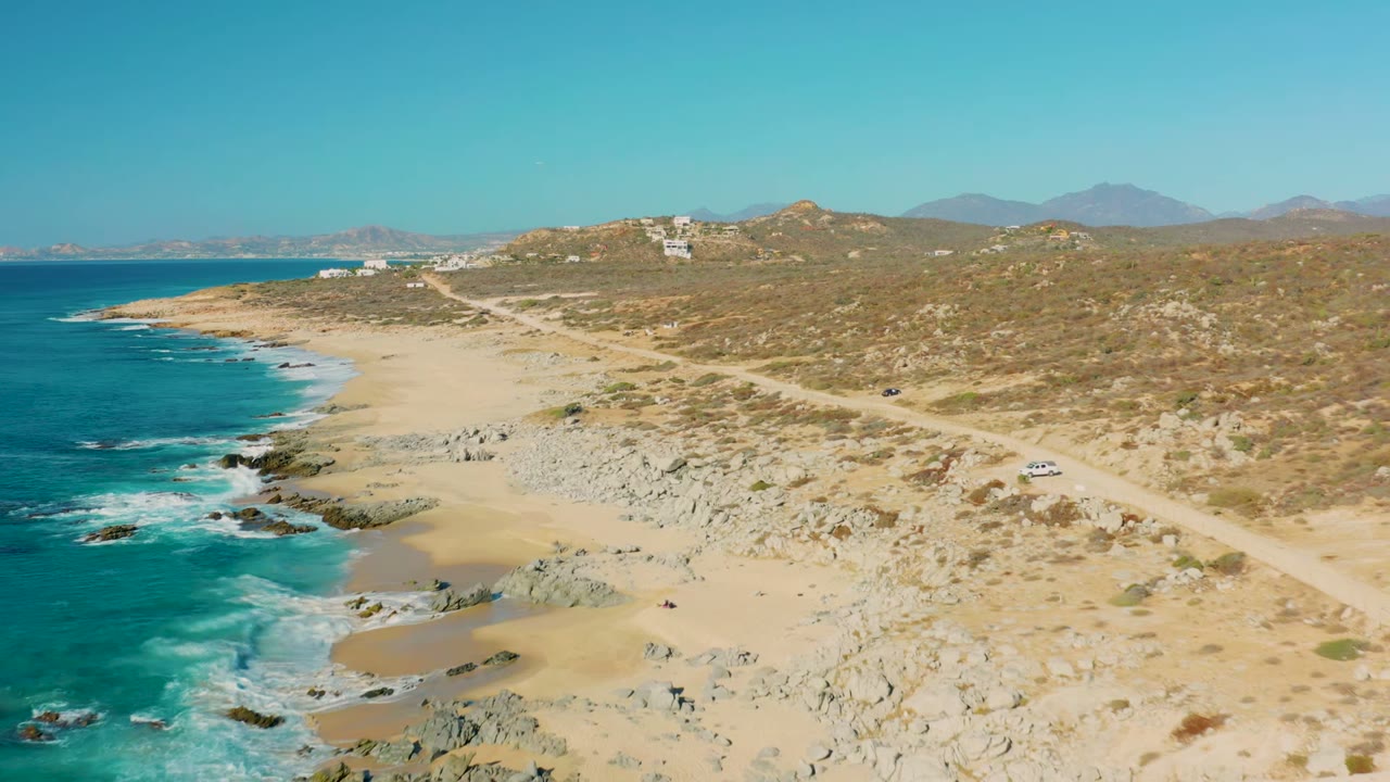 Aerial frame over La Fortuna, Baja California Sur, with turquoise ocean at left, rocky sand in the center, a dirt road on the right, a white car near the road, and no visible people.