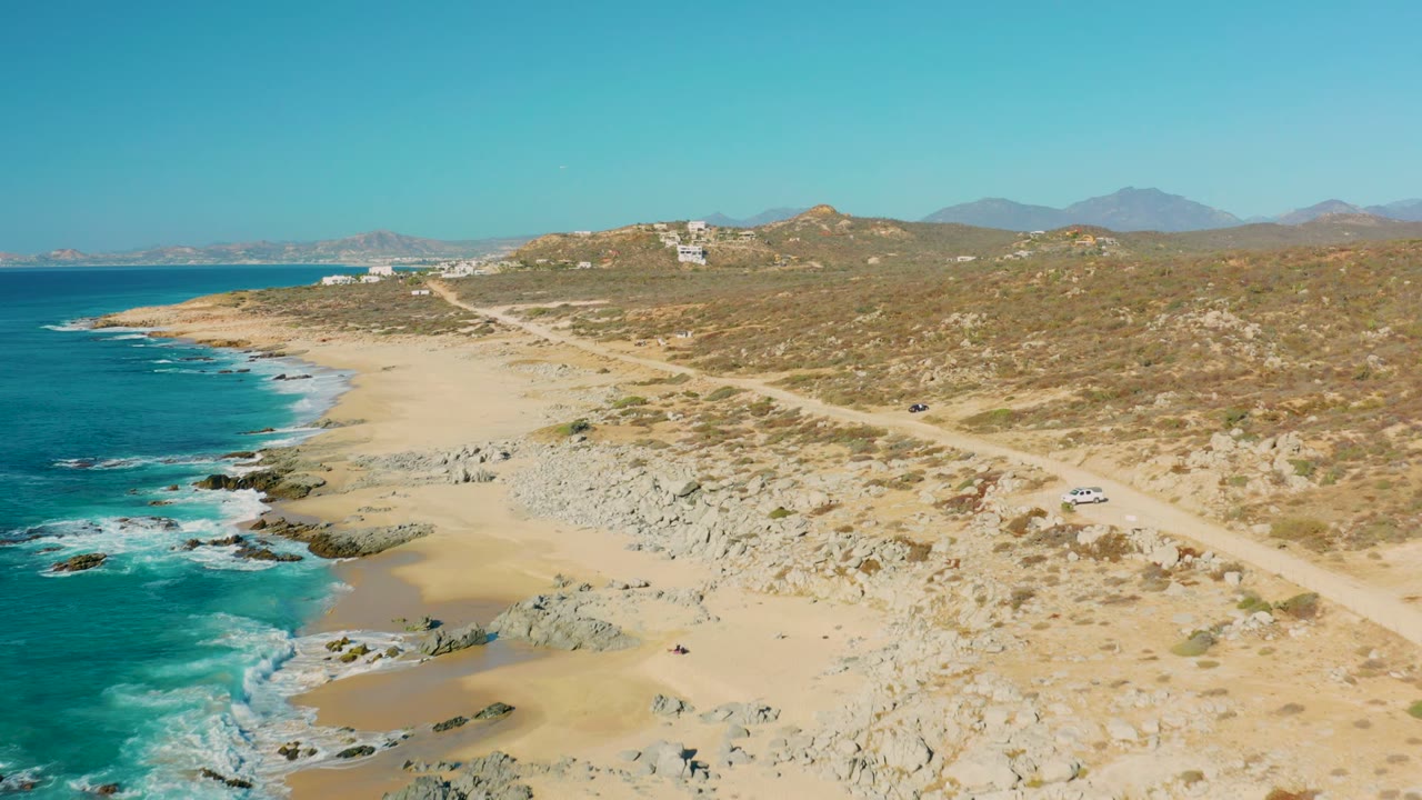 Aerial coastal view with waves, rocks, sand, dirt road, and a white car on the right.