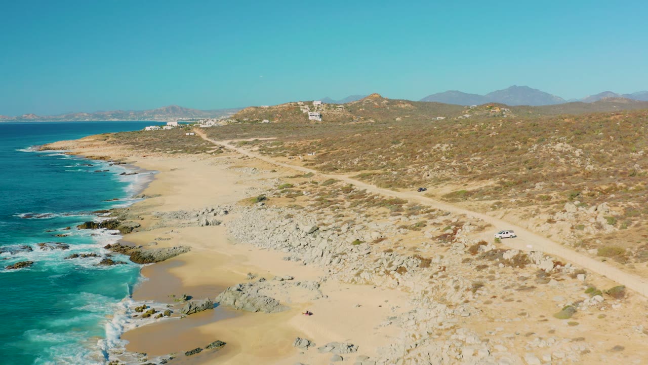 Drone-height view in La Fortuna with blue ocean along the left edge, pale beach and rock fields below, a winding dirt road through dry scrub, a white car on the right, and no visible people.