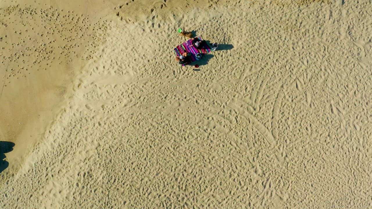 Overhead frame from La Fortuna, Baja California Sur, Mexico, showing a person on a striped blanket near the top of pale sand marked by footprints and a long shadow.
