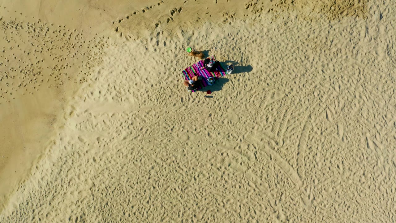 At 0:02, the La Fortuna beach frame keeps the person and colorful blanket near the upper right, with diagonal tracks and scattered footprints visible across the sunlit sand.
