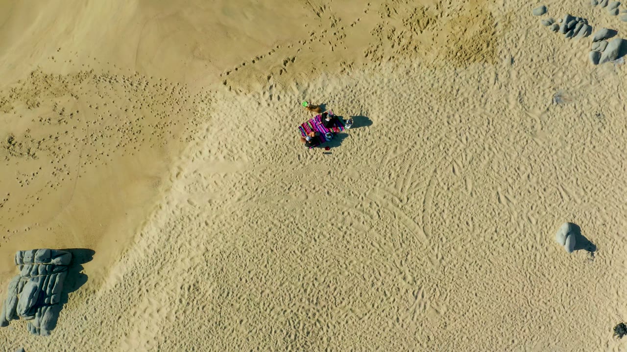 Overhead frame from La Fortuna, Baja California Sur, Mexico showing a purple dune buggy near the upper center of pale sand, with rock clusters at the lower left and lower right.