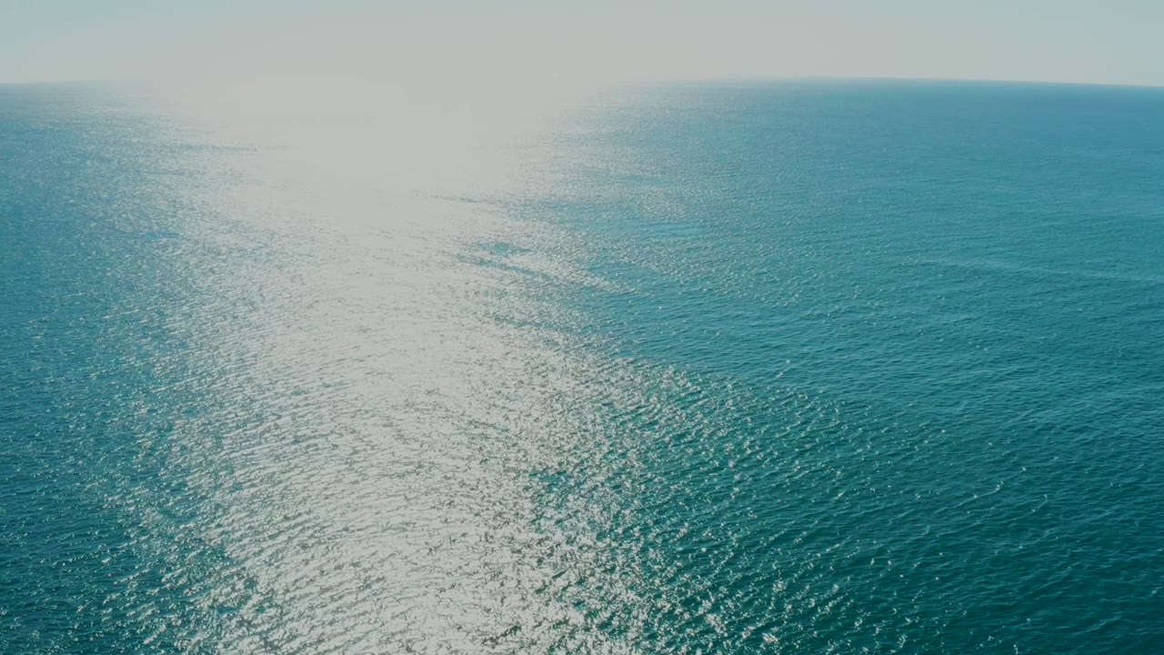 Wide aerial view over turquoise ocean at La Fortuna, Baja California Sur, with bright sun reflection on the left side of the water, distant coastline on the horizon, and no visible people.