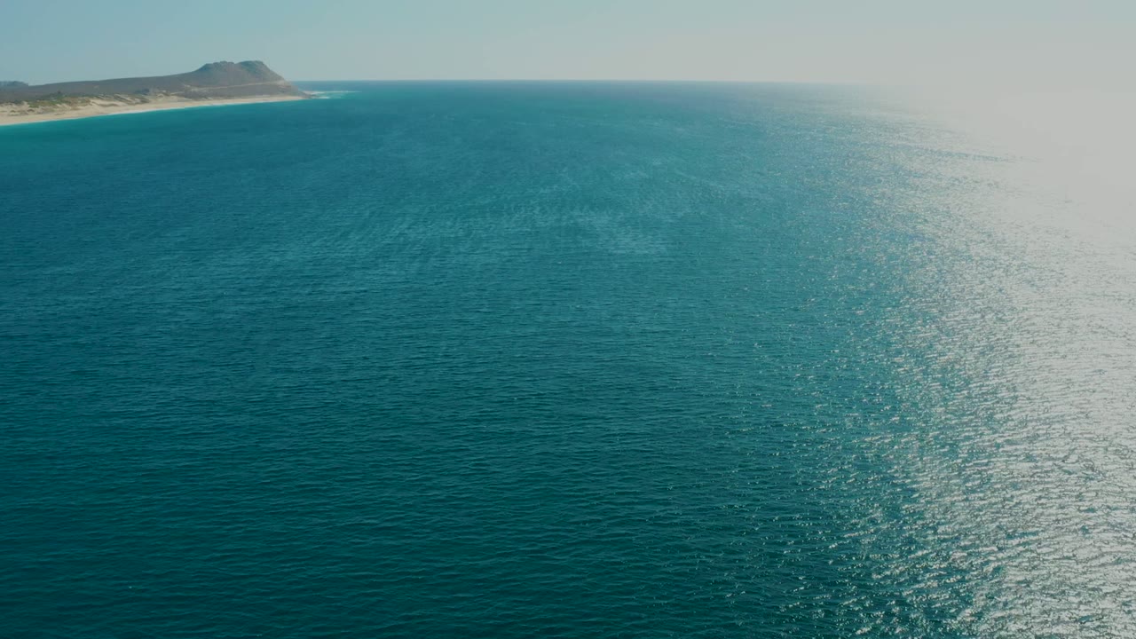 Aerial view of turquoise sea at La Fortuna, Baja California Sur, with a distant beach and low hill along the upper left horizon, sunlit water texture, and no visible people.