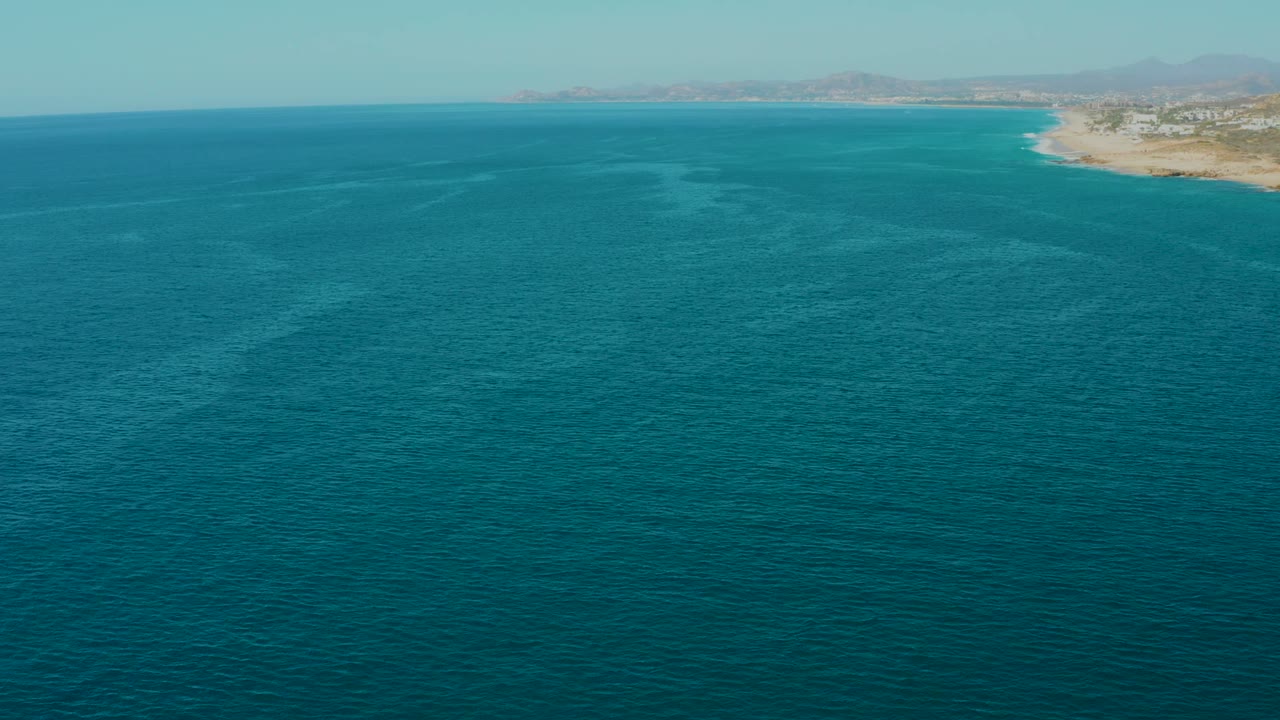 Aerial ocean frame in La Fortuna, Baja California Sur, Mexico with rippled blue water filling the view and only a narrow strip of coastline and mountains near the upper right, with no visible people.