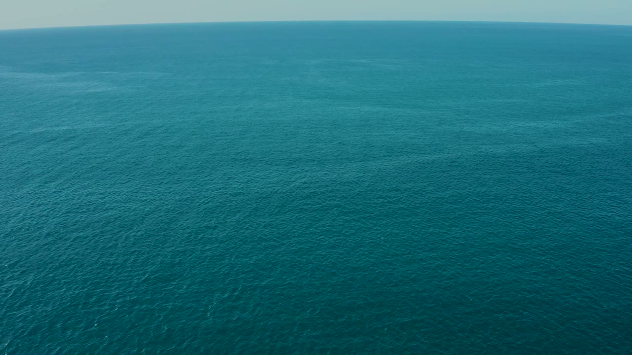 Wide aerial frame over blue-green ocean at La Fortuna, Baja California Sur, with fine surface ripples, a pale sky band, and no visible people.