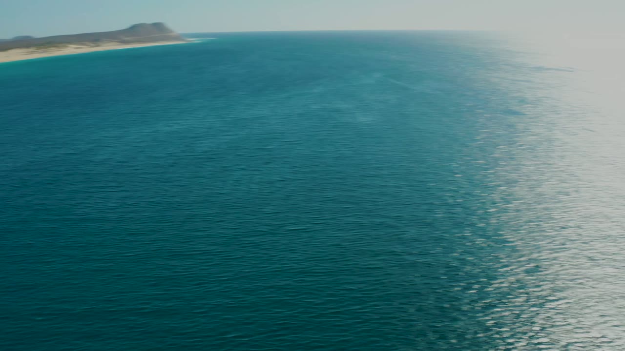 Aerial view near La Fortuna, Baja California Sur, with a small sandy island edge at the upper left, blue ocean filling the frame, sun haze on the horizon, and no visible people.