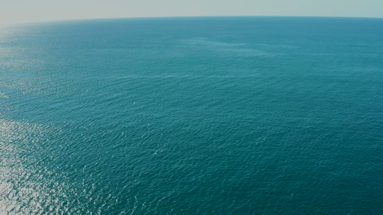 Wide aerial frame over open turquoise-blue water near La Fortuna, Baja California Sur, showing low ripples, a small bright reflection patch, a faint horizon, and no visible people.