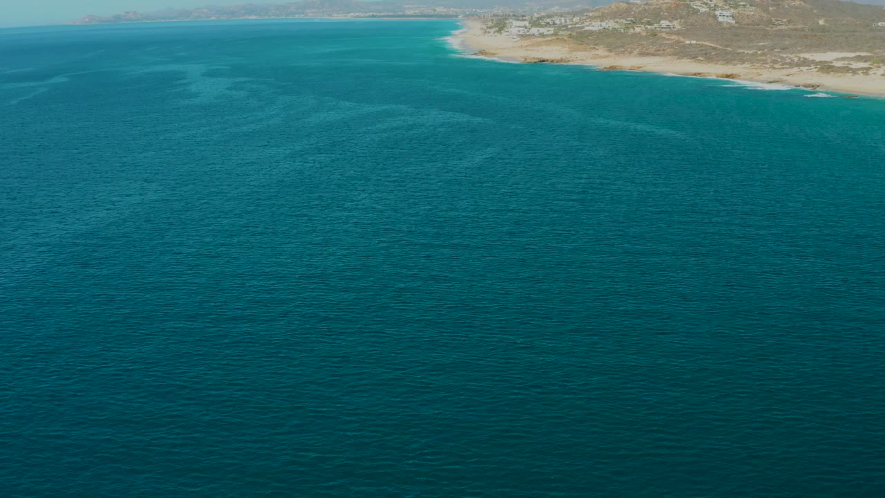 Opening aerial frame over La Fortuna, Baja California Sur, with turquoise ocean filling most of the view, sandy shore and dry hills along the top edge, and no visible people.