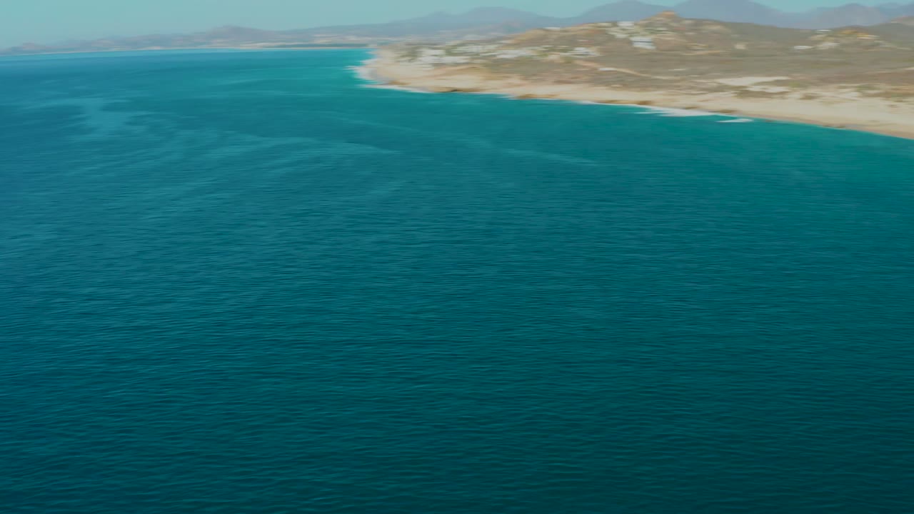 Wide aerial coastline with blue ocean, pale beach, hills, and distant mountains.