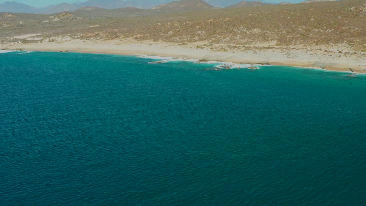 The La Fortuna shoreline runs across the upper frame, with textured turquoise water in front, tan beach and low hills behind it, distant mountains, and no visible people.