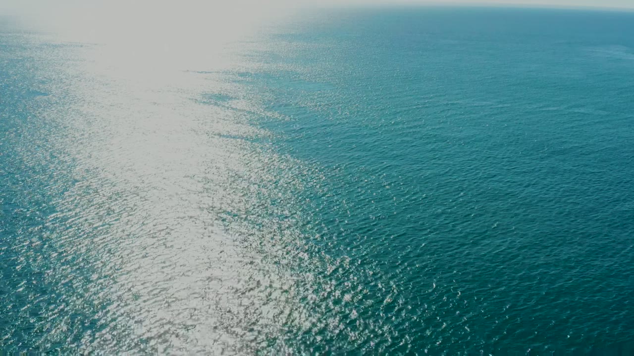 Wide aerial frame over the ocean near La Fortuna, Baja California Sur, with bright sun reflection on the left side, deep blue rippled water, a distant horizon, and no visible people.